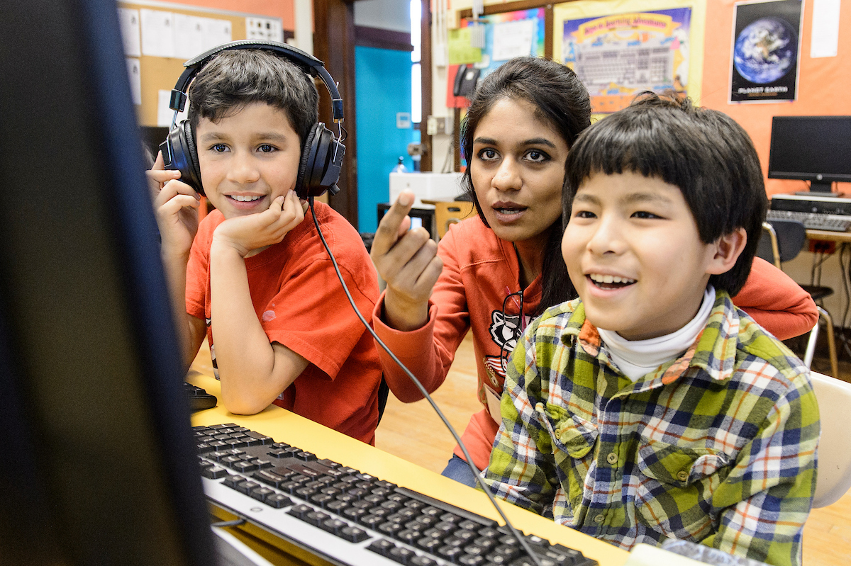 Two elementary students and a club facilitator look towards a computer screen. The students smile as the club facilitator provides guidance.]